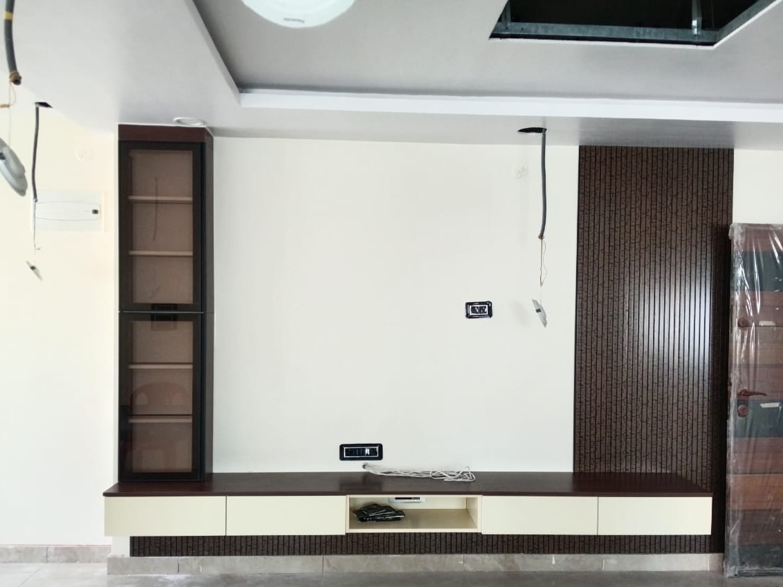 Living-room TV wall during installation: a long wall-hung console with cream drawers and an open niche, a tall glass display unit in a black frame on the left, and a full-height espresso fluted panel on the right.