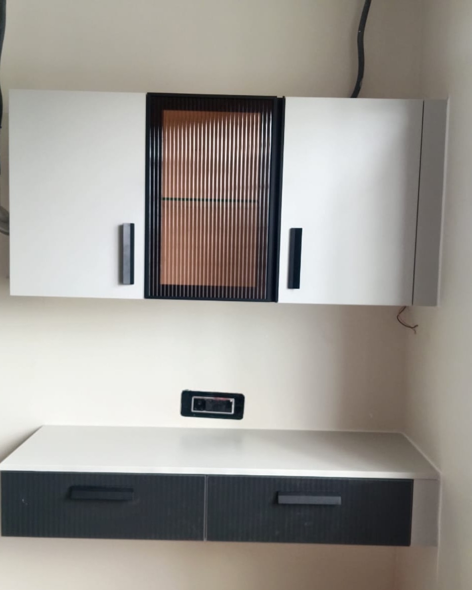 Study wall during installation: a white overhead cabinet with a central reeded-glass shutter in a black frame, above a floating white desk with two charcoal fluted drawers.